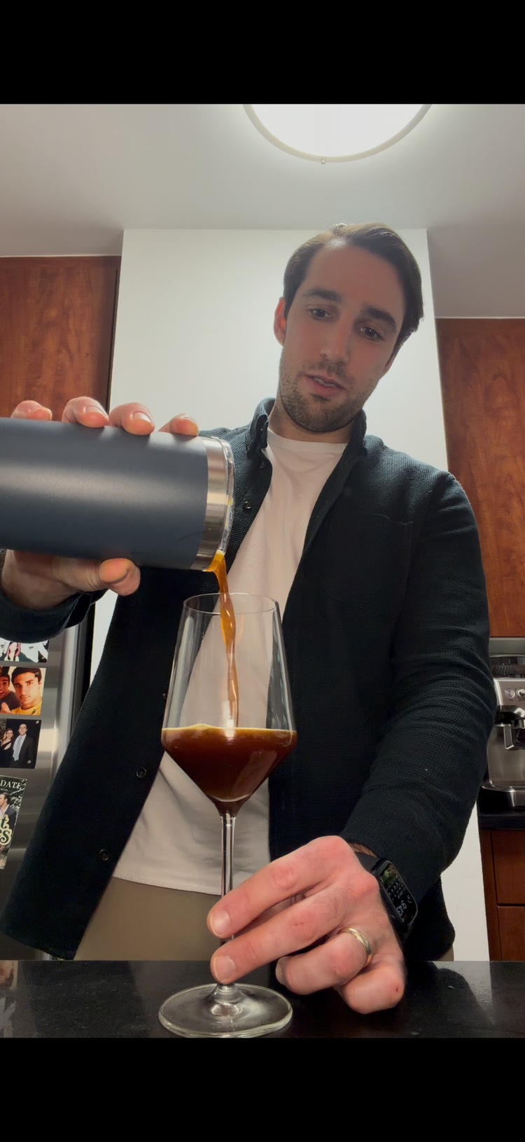 Person pouring an espresso martini from a cocktail shaker into a glass in a kitchen setting.
