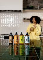 Row of four Motif syrup bottles on a green tiled counter with a woman in a yellow sweater enjoying a drink in the blurred background.