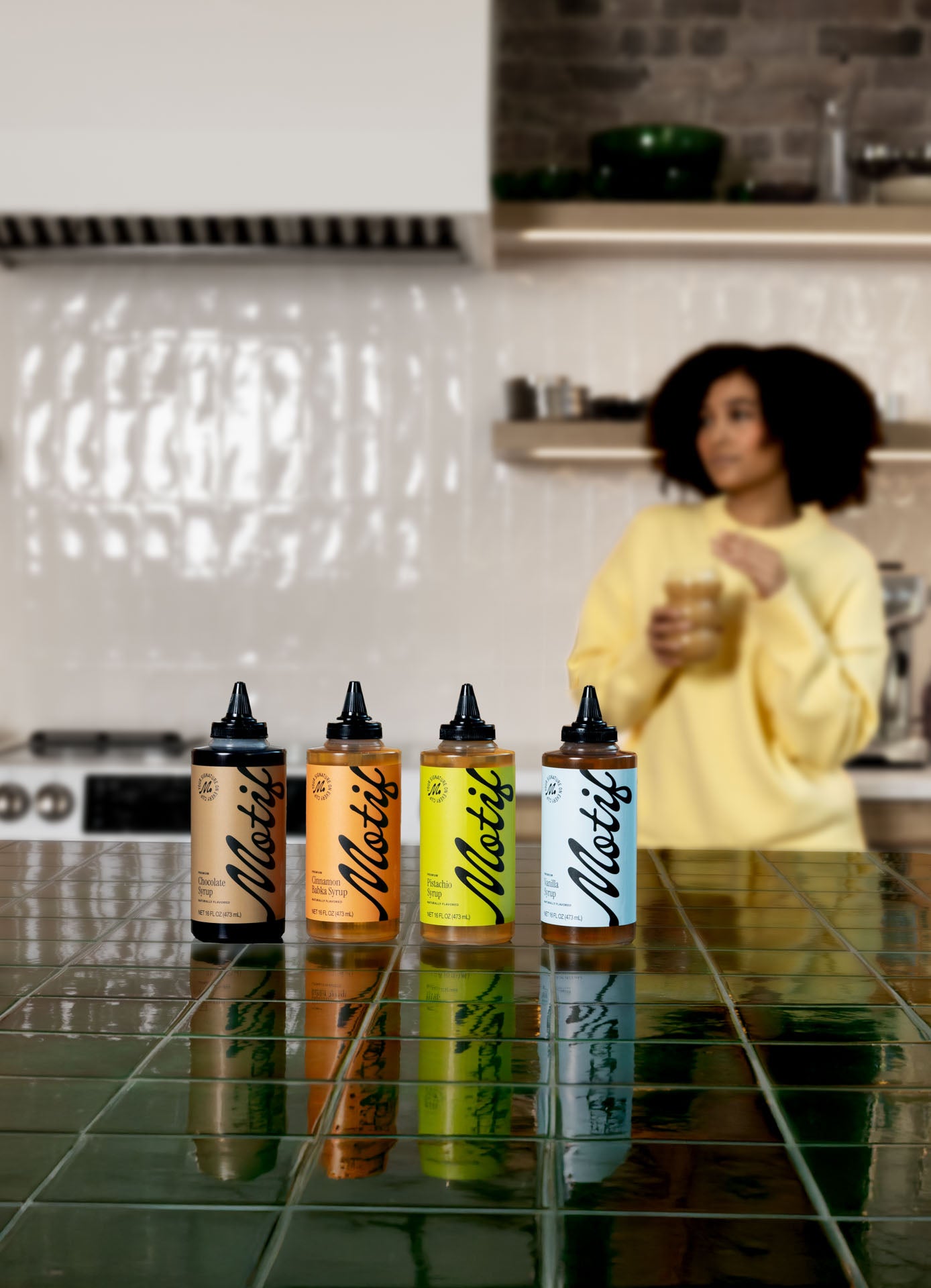 Row of four Motif syrup bottles on a green tiled counter with a woman in a yellow sweater enjoying a drink in the blurred background.