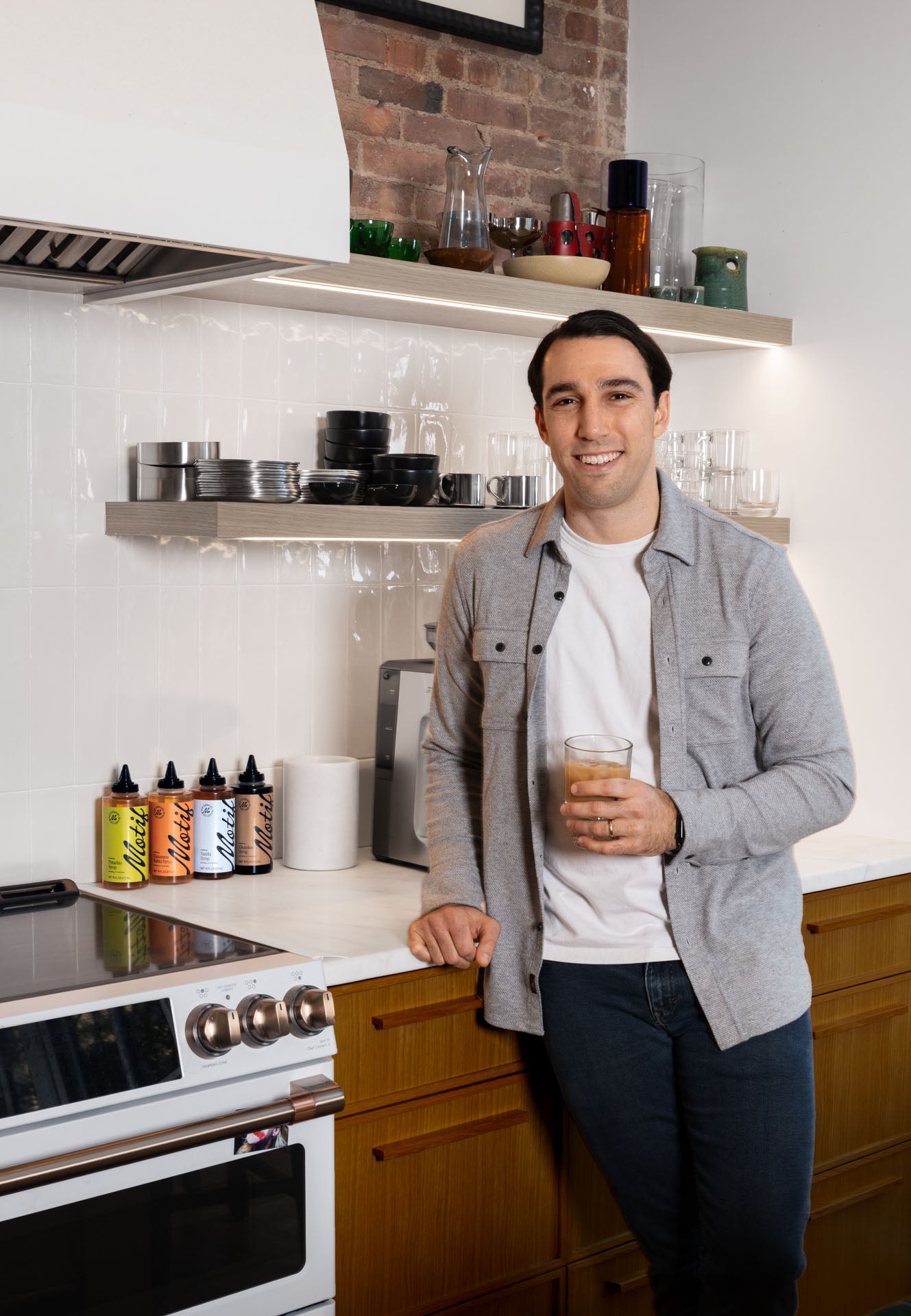 Founder Michael Zabar leaning against a kitchen counter holding an iced coffee next to a row of Motif syrups.