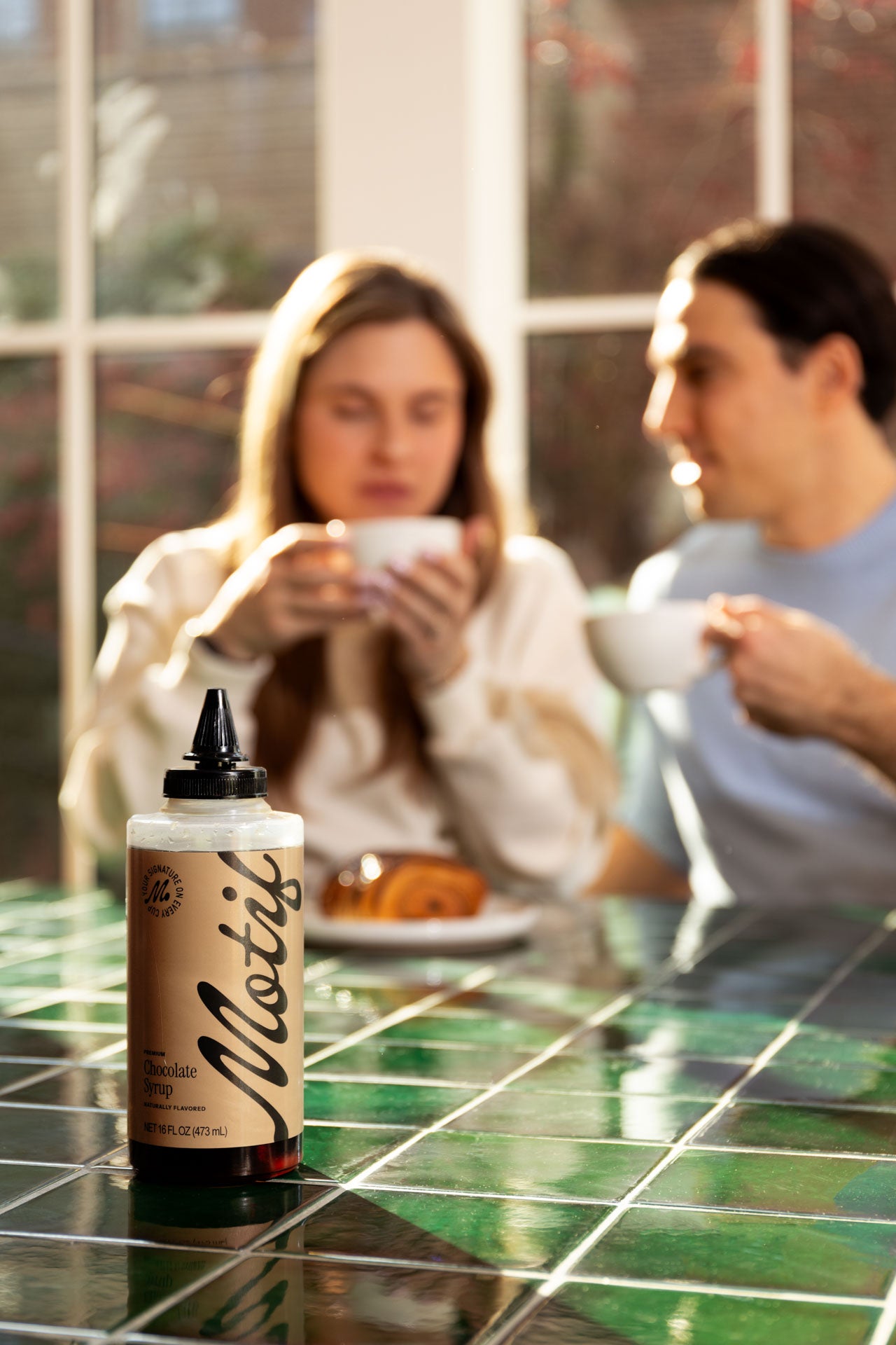 Bottle of Chocolate Syrup in sharp focus on a table, with a couple drinking coffee in the blurred background.