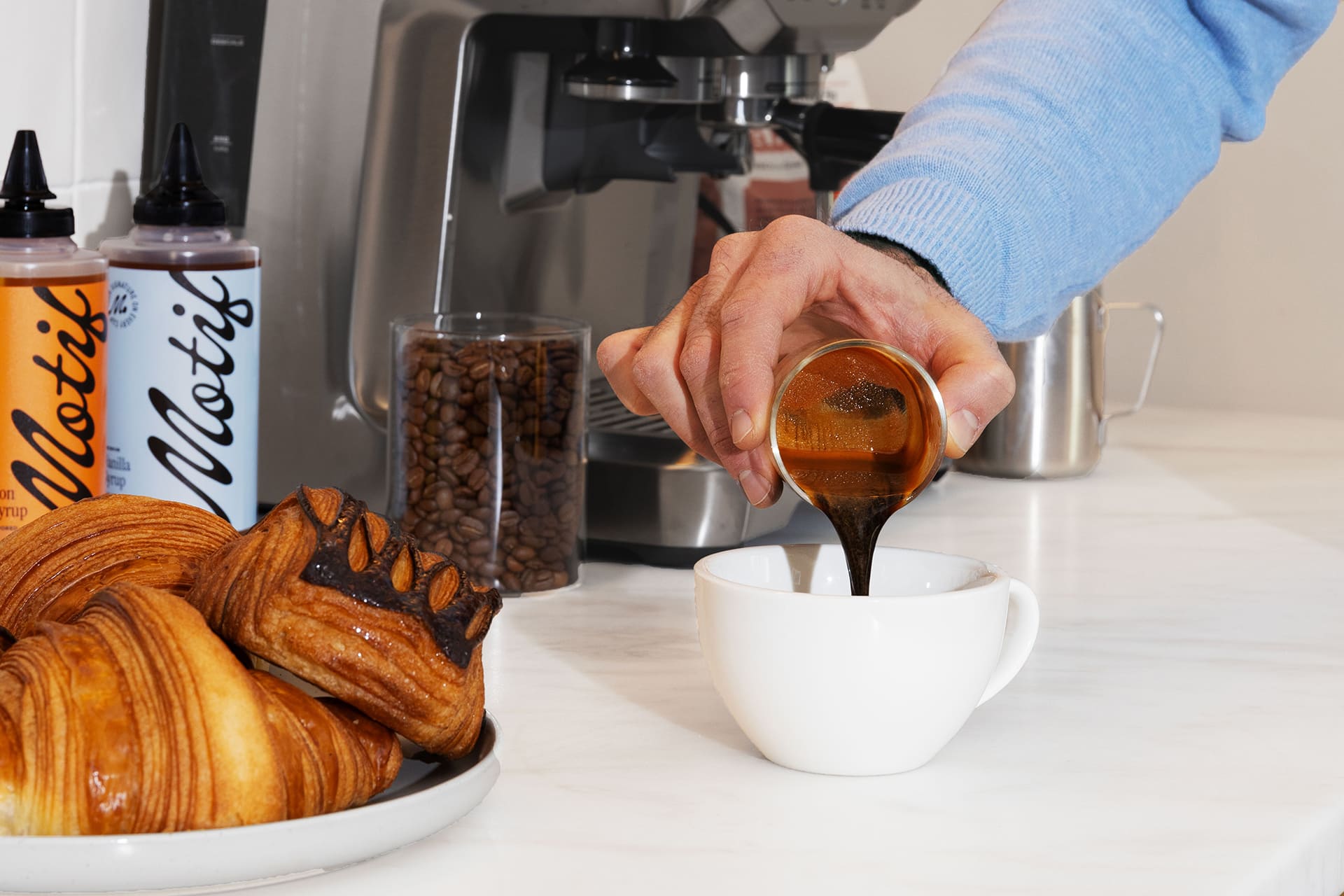 Hand pouring a shot of espresso into a white cup next to a plate of croissants.