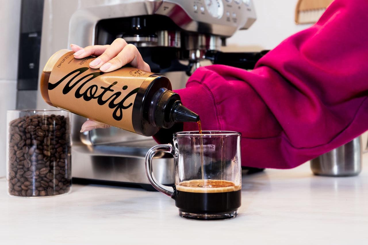 Step 2: Person squeezing Motif syrup from a bottle into a clear glass coffee mug.