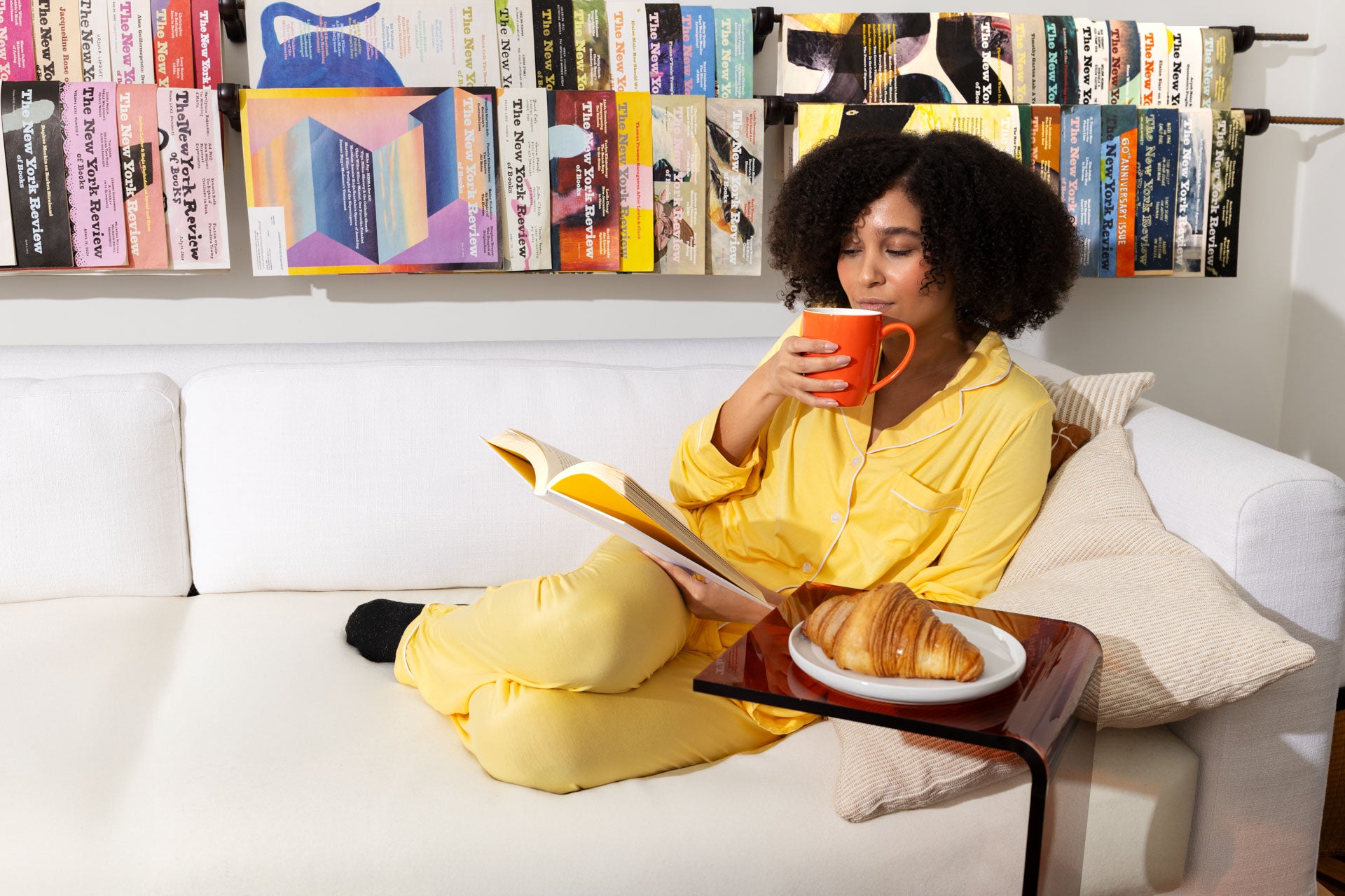 Woman in yellow pajamas relaxing on a white sofa, sipping coffee and reading a book next to a fresh croissant.