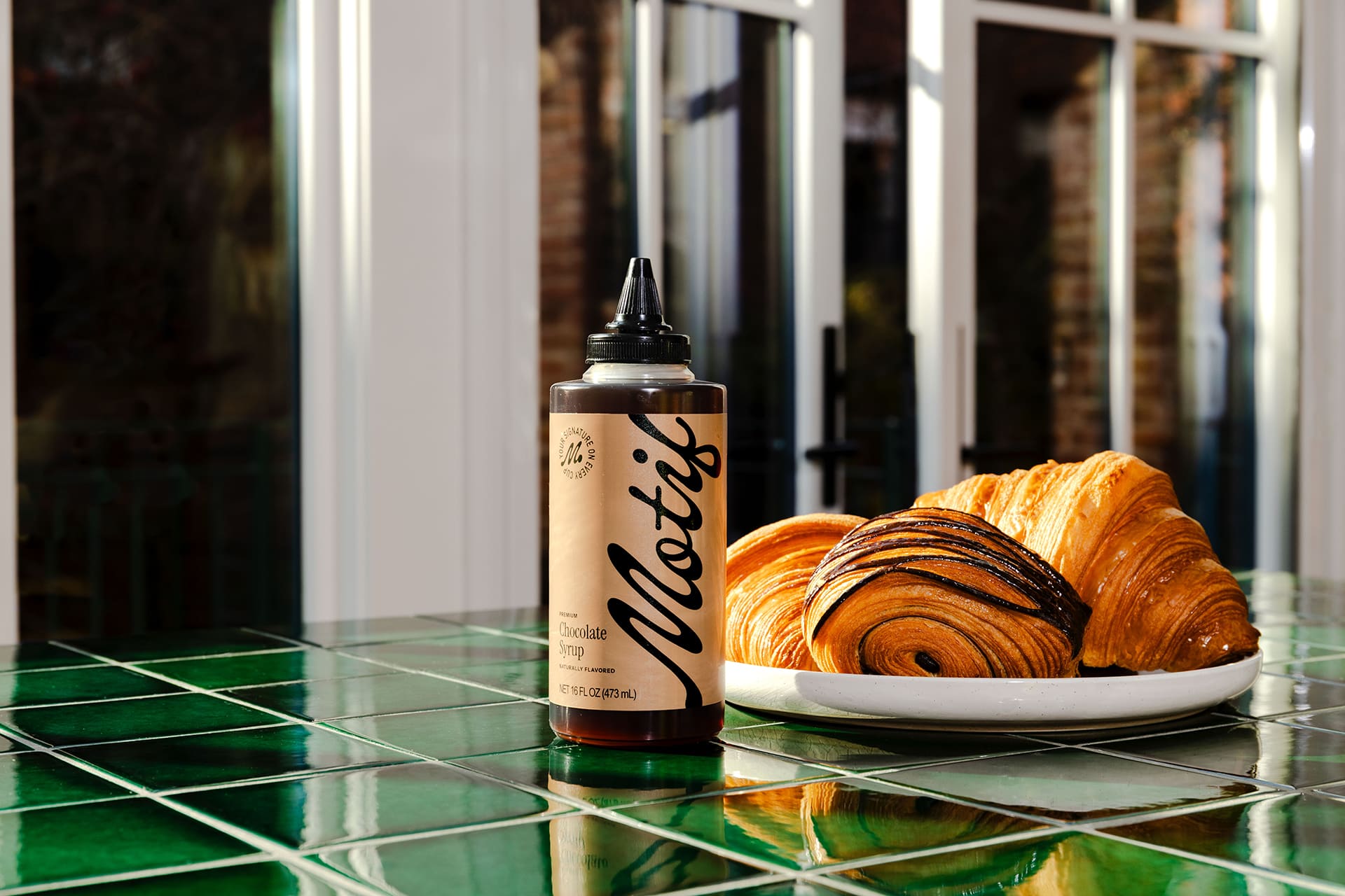 Bottle of Chocolate Syrup displayed on a green tiled counter next to a plate of chocolate croissants.