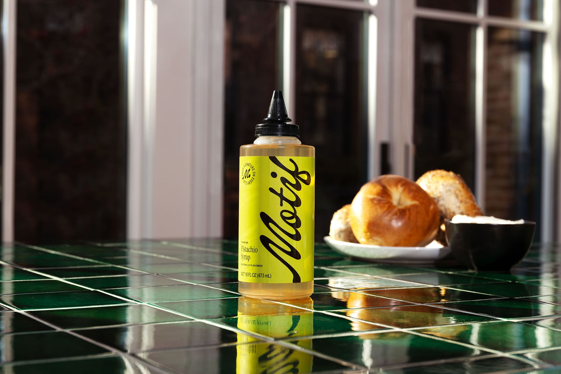 Bottle of Pistachio Syrup on a green tiled counter next to a plate of fresh bagels.