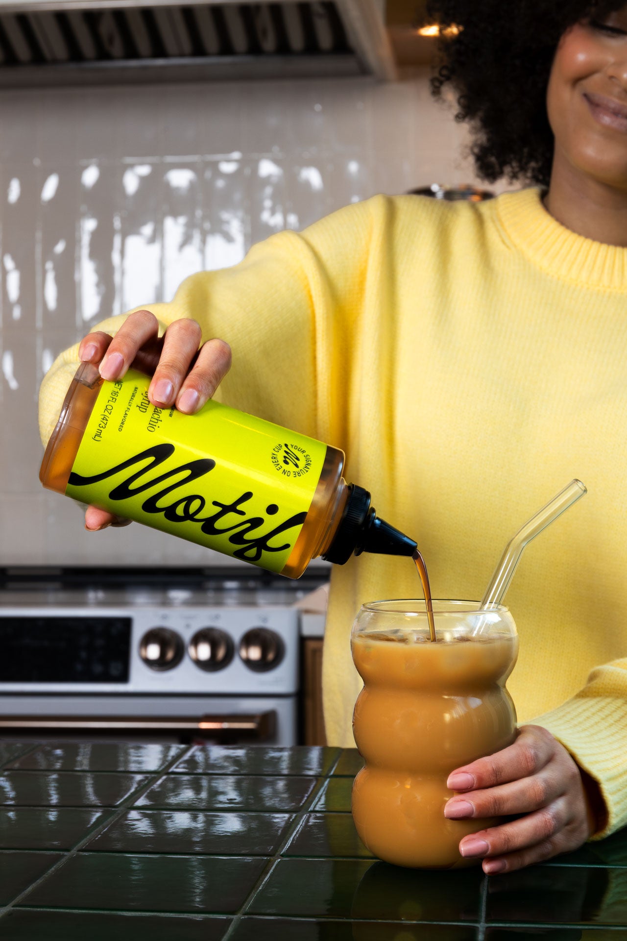 Woman in a yellow sweater pouring Pistachio Syrup into an iced coffee using the bottle's precision cap.